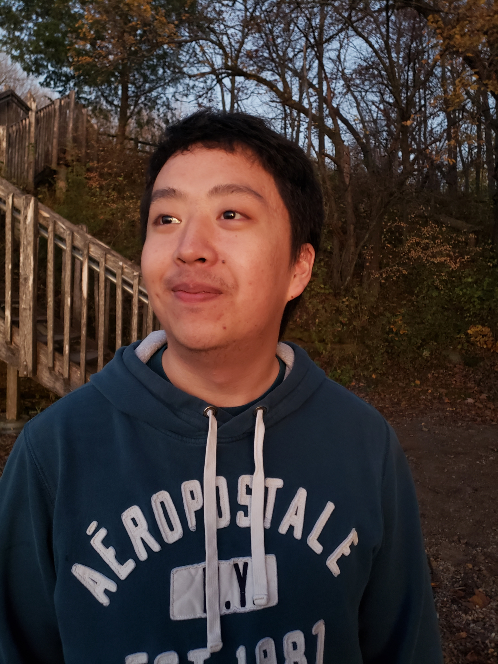 Smiling Hmong man wearing a green aeropostale sweatshirt while standing in front of a forest